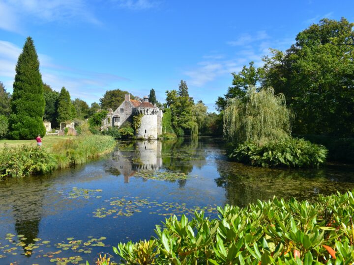 A silhouette of a woman drawing a British medieval castle and its reflection in water. Romantic gardens with panoramic views of a moated fortress and a Victorian country mansion all in a wooded estate
